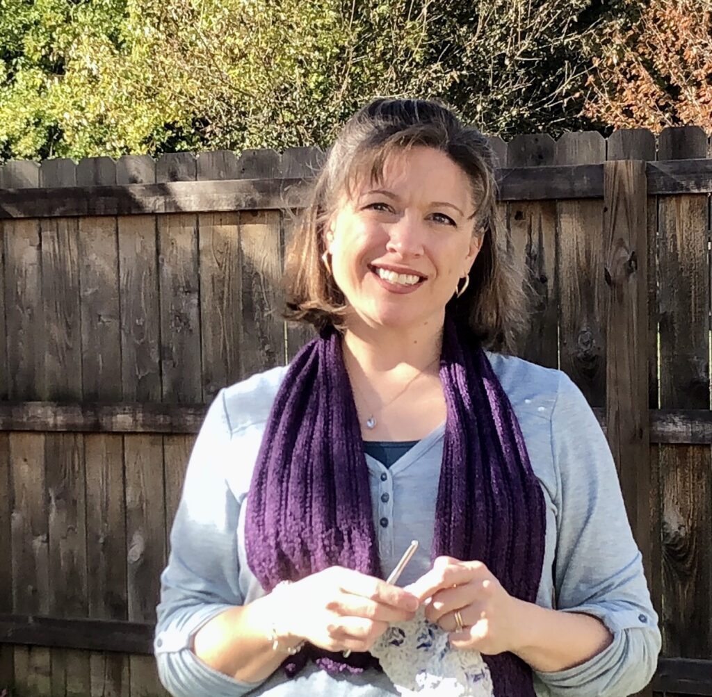 photo of Jenn standing in front of a fence wearing a light blue shirt and purple scarf holding crochet.