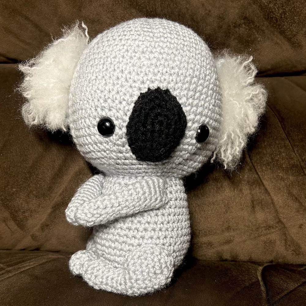 gray crocheted koala with black nose and eyes and fuzzy white ears sitting on a dark brown chair.