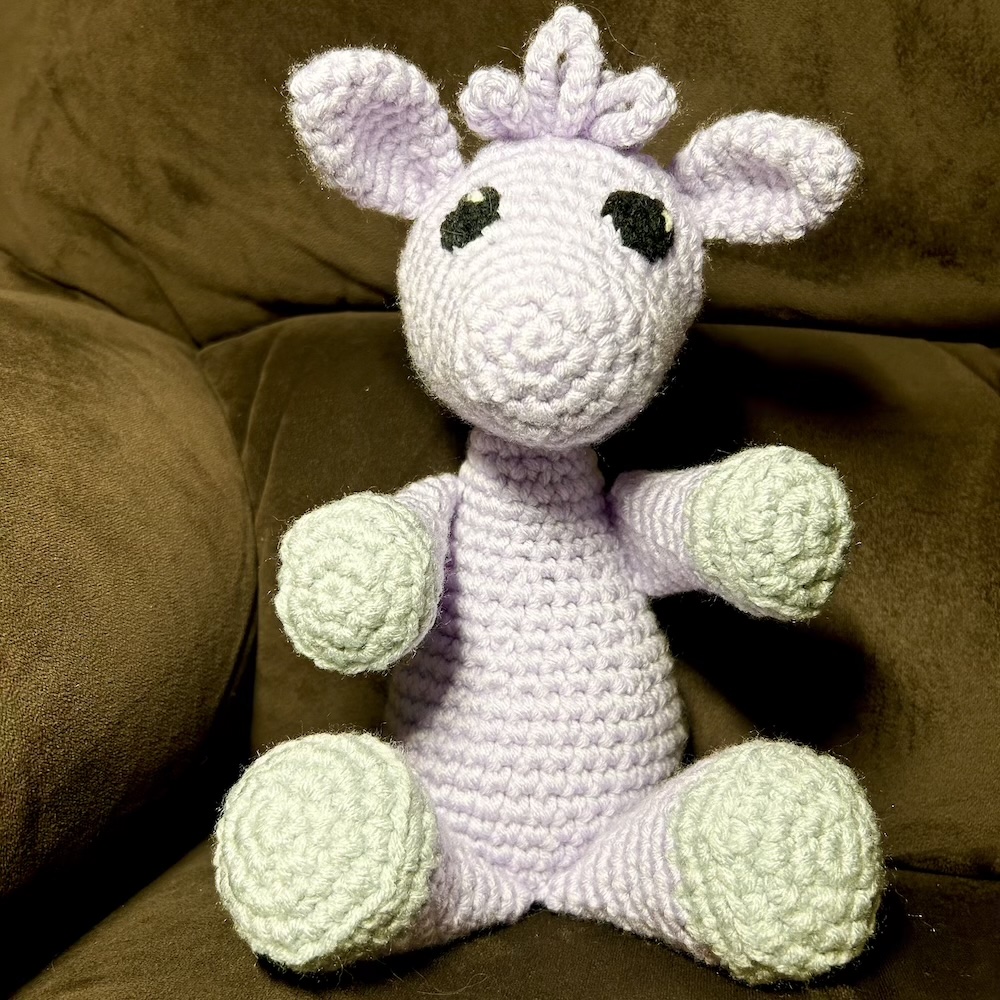 lavender crocheted llama with gray hooves sitting on a brown chair.