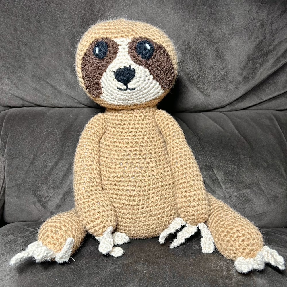 tan and dark brown crocheted sloth sitting on a dark brown chair.