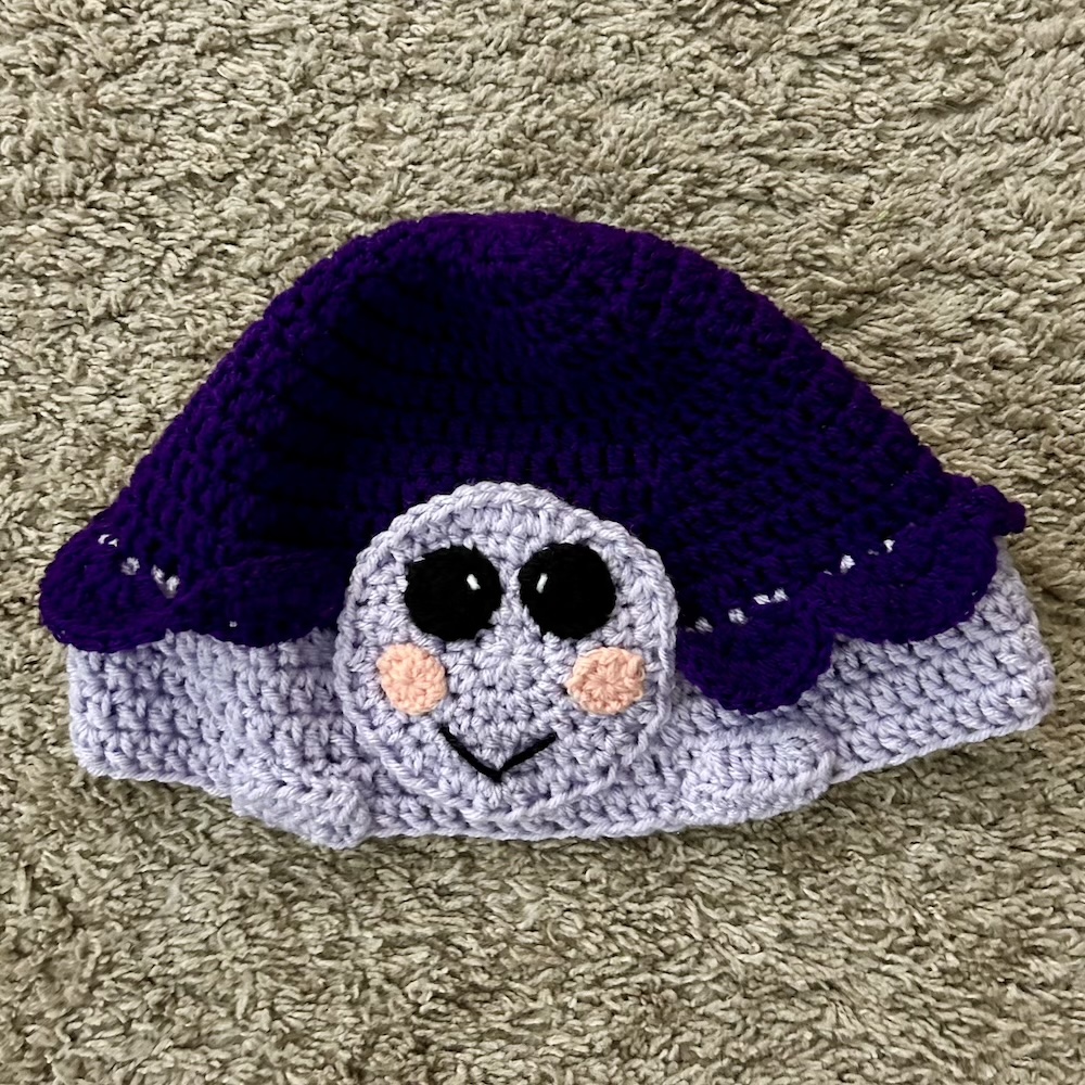 dark and light purple crocheted turtle hat on a tan background.