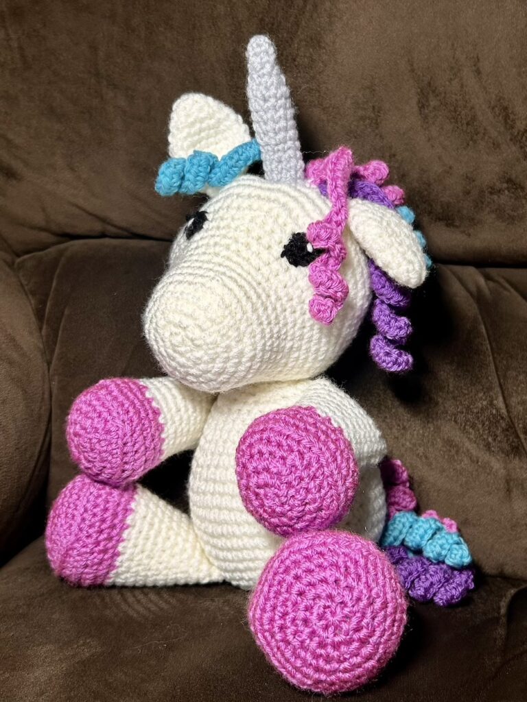 white crochet unicorn with pink hooves, pink/purple/turquiose curly hair and a gray horn sitting on a dark brown chair.