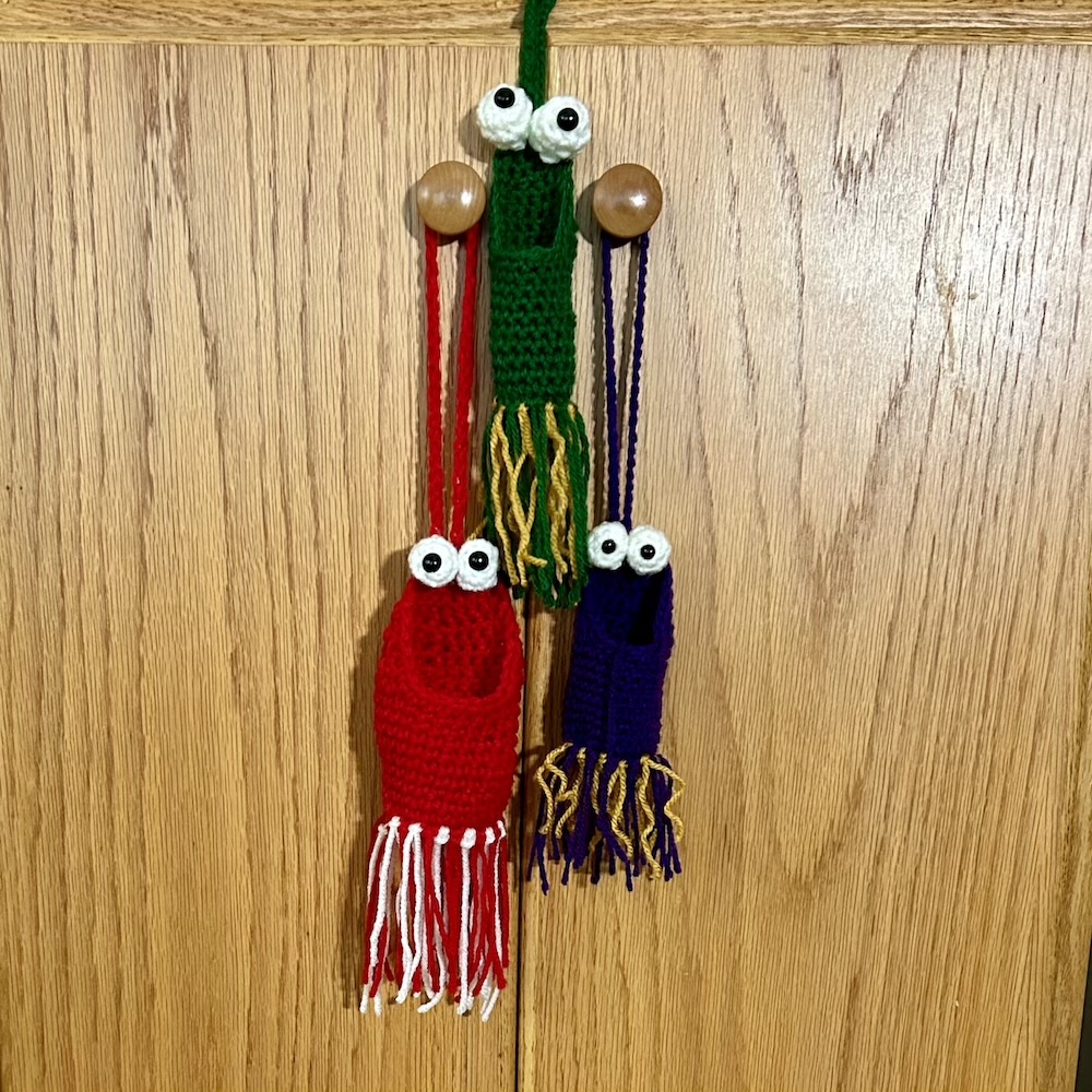 red, purple and green crocheted "Yip Yips" handing on a wooden cabinet.