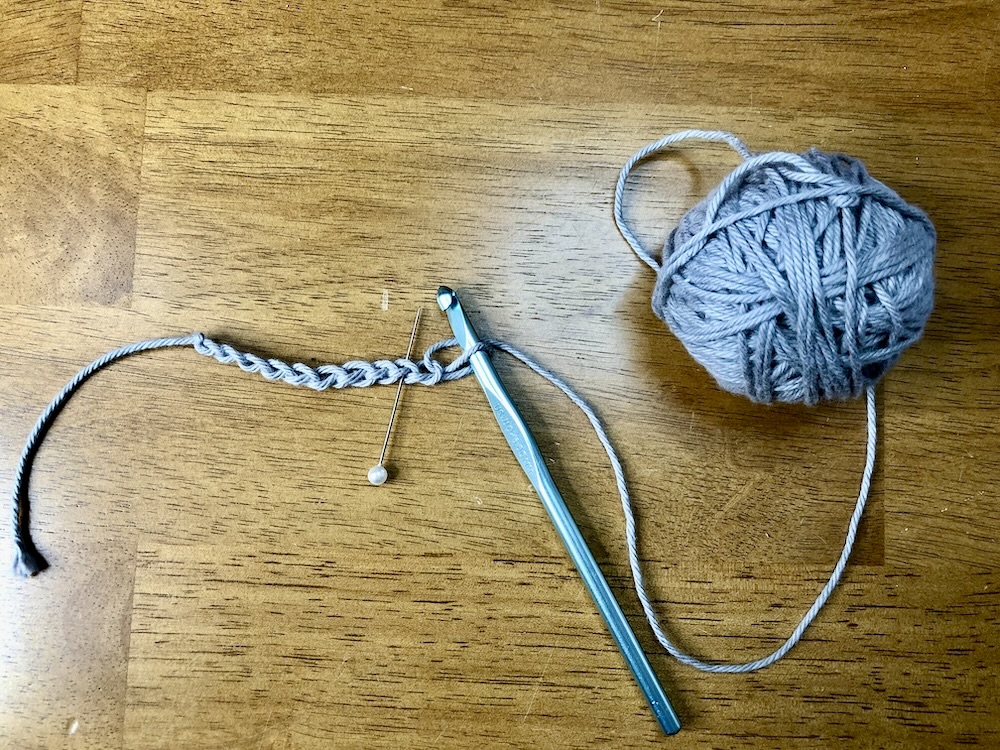 ball of gray yarn with a crochet hook and a foundation chain with a pin inserted on a wooden background.
