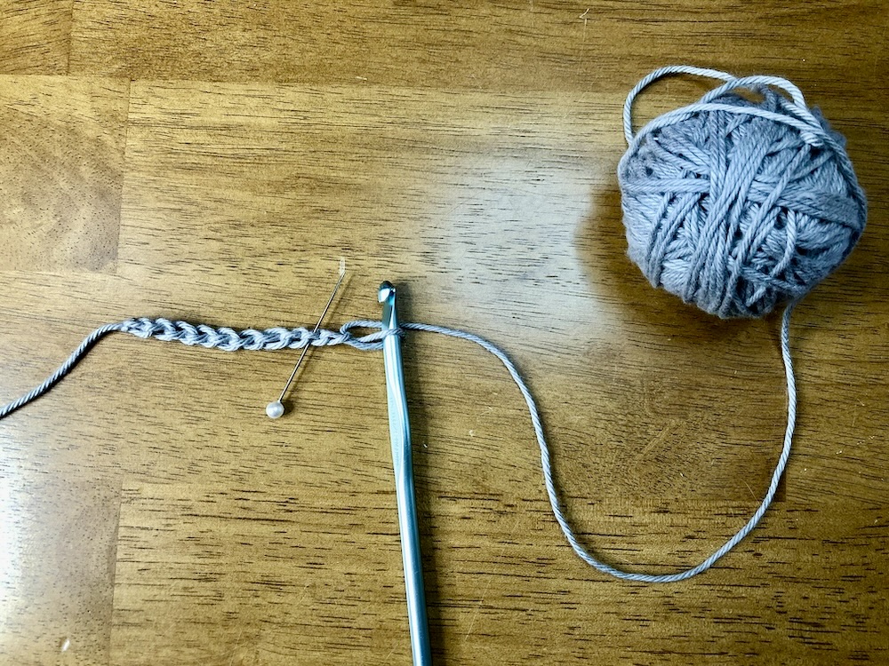 ball of gray yarn with a crochet hook and a foundation chain with a pin inserted on a wooden background.