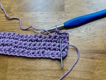 crocheted strip with purple yarn and a needle showing stitch placement on a wooden background.