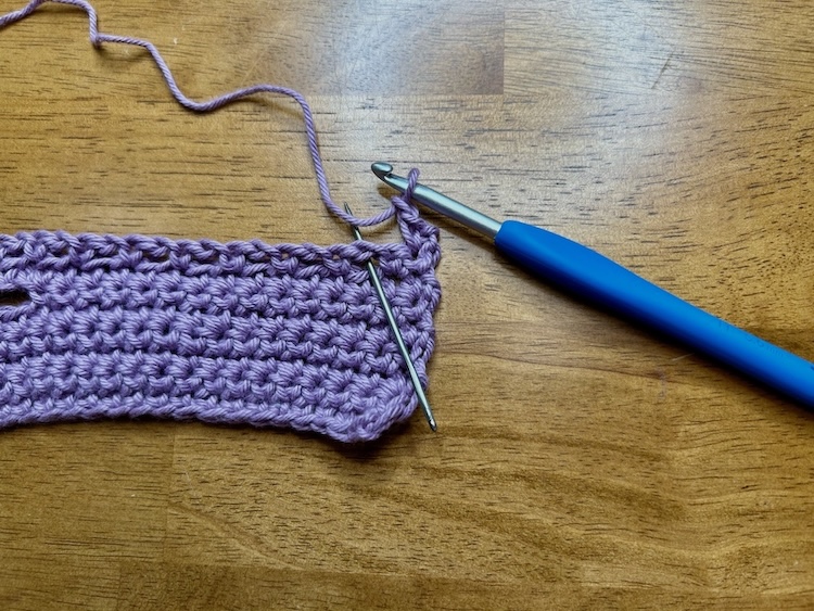 crocheted strip with purple yarn and a needle showing stitch placement on a wooden background.