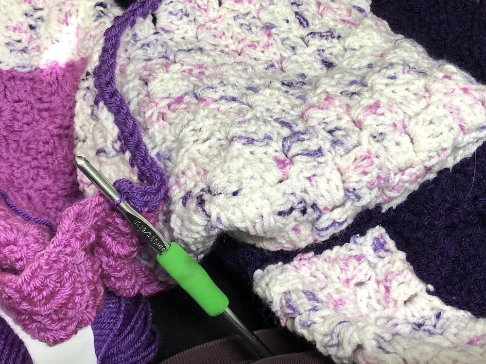 close up of white, pink and purple crocheted blanket with green handled crochet hook.